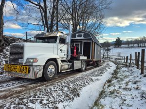 Delivery of a lofted barn-style storage shed with porch, two windows, and entry door on a trailer along a snowy rural country road with fields and mountains in the background.
