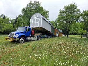 Delivery truck unloading multiple storage sheds into wildflower field with trees in rural setting.